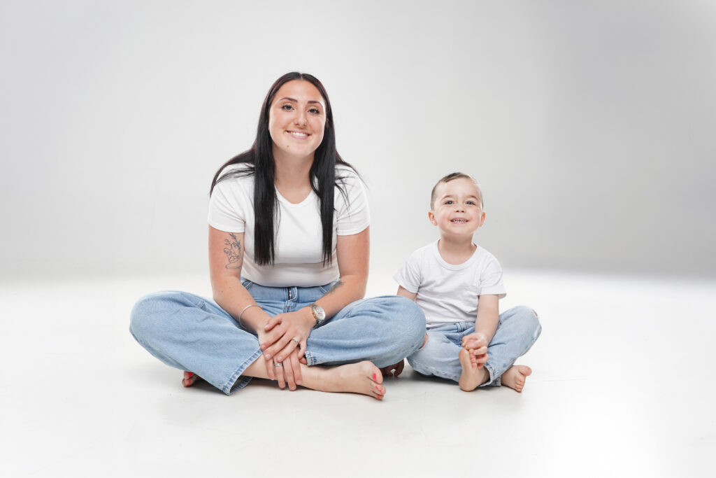 Portrait studio épuré d'une maman et son petit garçon assis en tailleur en t-shirts blancs et jeans.
