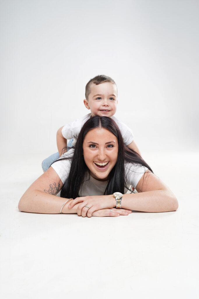 Moment de jeu capturé en studio avec un enfant sur le dos de sa maman allongée au sol, tous deux très souriants.